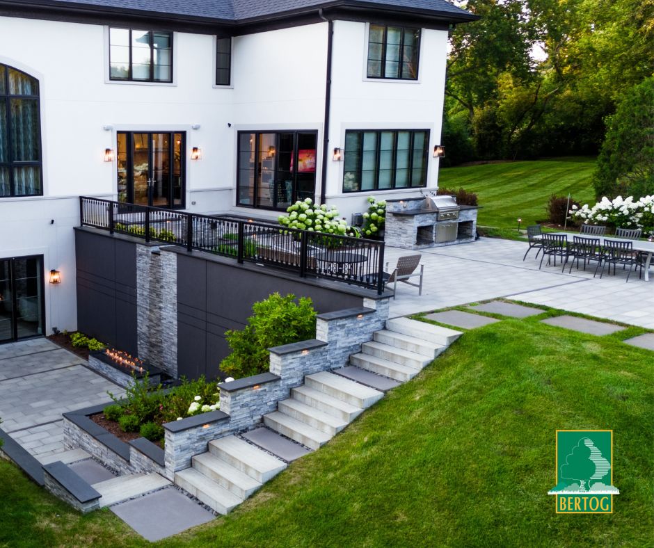 Two-level landscape with tiered retaining walls, natural stone steps, outdoor kitchen, and lower patio with linear fire table — designed by Green Grass Landscape.
